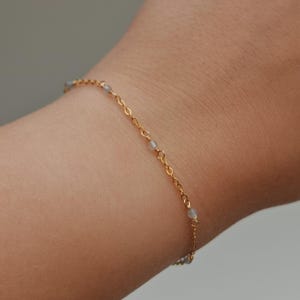 Peut inclure: Un bracelet doré délicat orné de petites perles bleu clair. Le bracelet est porté sur un poignet, mettant en valeur sa fine chaîne et son design subtil. Les perles sont espacées uniformément.