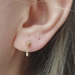 May include: A gold-colored earring with a small, round bead at the top and a dangling, clear, teardrop-shaped bead below. The earring is shown in the context of a person's ear.