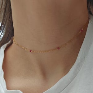 May include: A delicate gold-colored choker necklace with small, round, red beads evenly spaced along the chain. The necklace is worn against a white t-shirt.