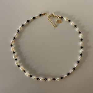 Puede incluir: Un delicado collar con perlas blancas de forma irregular intercaladas con peque&ntilde;as cuentas negras. El collar tiene un cierre y una cadena ajustable de color dorado, con un peque&ntilde;o dije en forma de coraz&oacute;n.