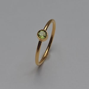 Op de afbeelding: Een dunne gouden ring met een enkele, ronde, gefacetteerde lichtgroene edelsteen in een eenvoudige zetting. Het metaal heeft een gepolijste, reflecterende afwerking en de steen is geplaatst aan de bovenkant van de ronde ringstructuur.