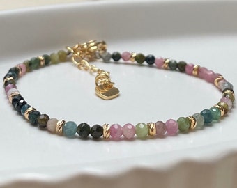 May include: A dainty bracelet featuring small, multi-coloured gemstone beads. The beads are a mix of pink, green, and blue, interspersed with gold accents. The bracelet has a heart-shaped charm attached.