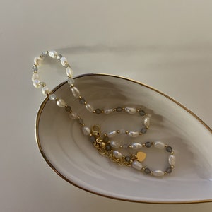 Puede incluir: Un collar de cuentas que alterna perlas de agua dulce blancas y pequeñas cuentas de piedra gris facetadas, acentuado con diminutas esferas de metal dorado. El collar incluye un cierre de langosta dorado, una cadena extensora y un pequeño dije de corazón.