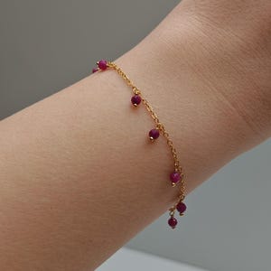 Könnte beinhalten: Ein zartes Armband aus goldfarbener Kette, verziert mit kleinen, runden, tiefroten Perlen. Die Perlen sind gleichmäßig entlang der Kette angeordnet und bilden ein schlichtes, aber elegantes Design. Das Armband wird am Handgelenk getragen.