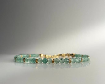 May include: A delicate bracelet featuring small, round, light green beads strung together with gold-colored accents. The bracelet is displayed on a white surface, with a soft, neutral background. The jewelry piece is designed for everyday wear.