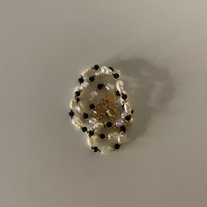 Puede incluir: Un collar de varias capas con perlas de agua dulce blancas y cuentas negras, con un cierre dorado. El collar se exhibe sobre una superficie blanca, mostrando su elegante dise&ntilde;o y colores contrastantes.