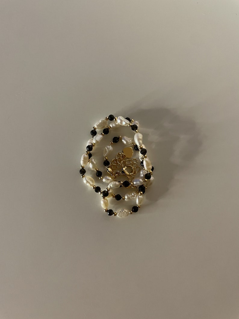 Puede incluir: Un collar de varias capas con perlas de agua dulce blancas y cuentas negras, con un cierre dorado. El collar se exhibe sobre una superficie blanca, mostrando su elegante dise&ntilde;o y colores contrastantes.
