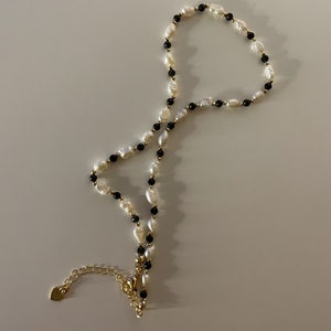 Puede incluir: Un delicado collar con perlas blancas de agua dulce y peque&ntilde;as cuentas negras, intercaladas con detalles dorados. El collar tiene una cadena y un cierre dorados. La joya se exhibe sobre una superficie blanca.