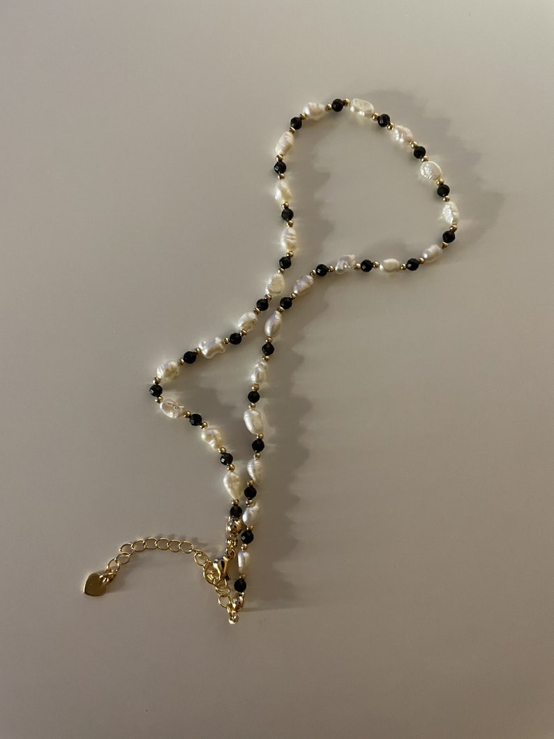 Puede incluir: Un delicado collar con perlas blancas de agua dulce y peque&ntilde;as cuentas negras, intercaladas con detalles dorados. El collar tiene una cadena y un cierre dorados. La joya se exhibe sobre una superficie blanca.