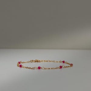 Può includere: Delicato braccialetto dorato con piccole perline rosa vivaci, distanziate lungo la catena. Il braccialetto ha una semplice chiusura ed è esposto su una superficie bianca, evidenziando il suo design elegante. Un gioiello.