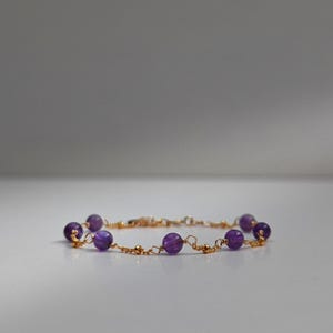 May include: A delicate bracelet featuring small, round, deep purple amethyst beads strung on a gold-coloured wire. The bracelet has a simple, elegant design, with the beads evenly spaced along the wire.