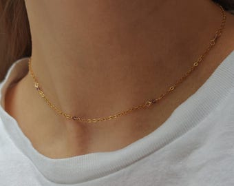 May include: A delicate gold chain necklace featuring small, evenly spaced purple gemstone accents. The thin, polished metal links form a simple, continuous strand that rests closely around the neck, highlighting the contrast between the gold and stones.