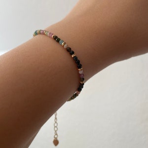 Puede incluir: Un brazalete delicado con una cadena dorada y cuentas de colores. Las cuentas son una mezcla de rosa, verde y negro.
