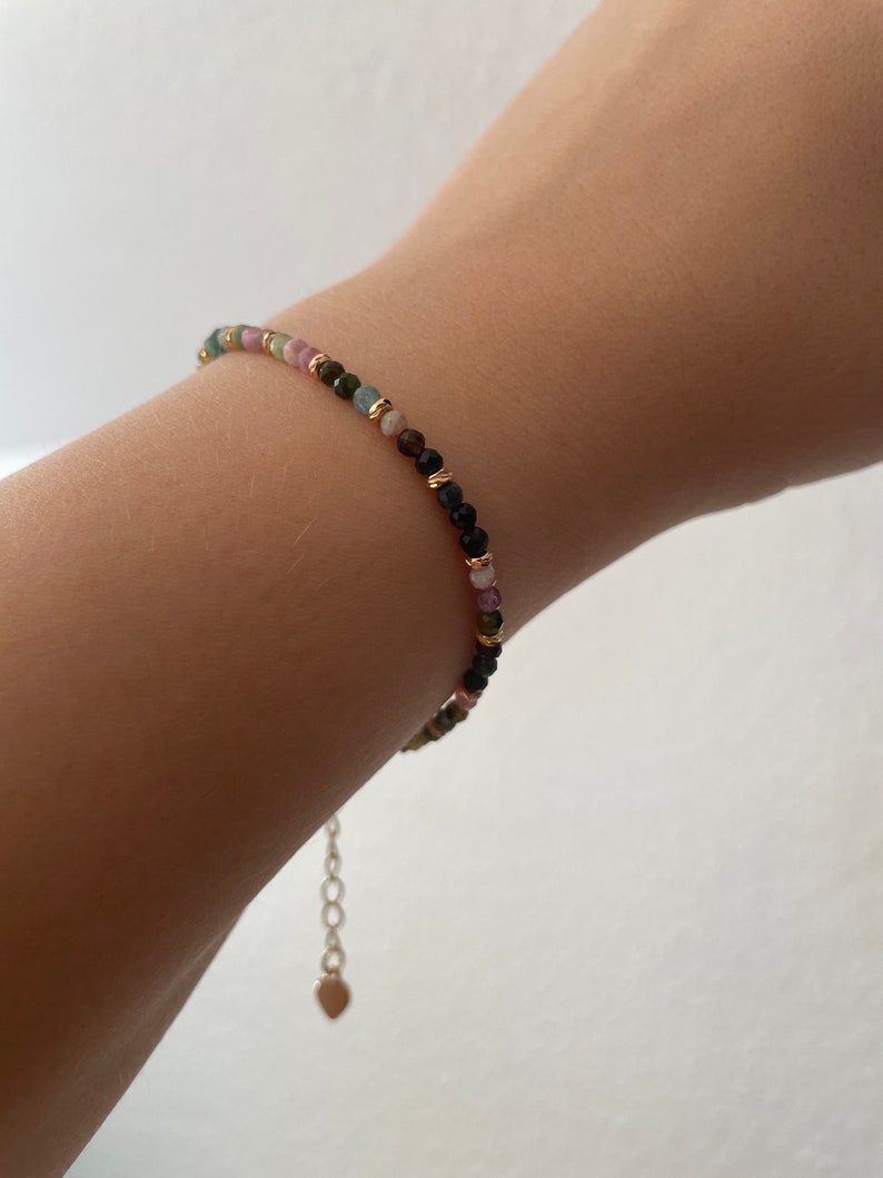 Puede incluir: Un brazalete delicado con una cadena dorada y cuentas de colores. Las cuentas son una mezcla de rosa, verde y negro.