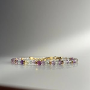 May include: A delicate bracelet made with small, faceted beads in shades of purple, green, and clear. The beads are strung on a gold chain.