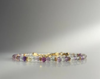 May include: A delicate bracelet made with small, faceted beads in shades of purple, green, and clear. The beads are strung on a gold chain.