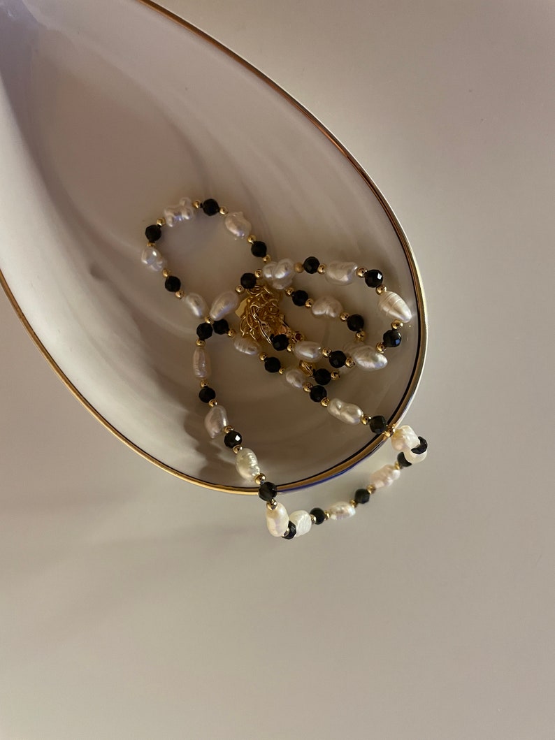 Puede incluir: Un delicado collar con una combinaci&oacute;n de perlas blancas y cuentas negras, intercaladas con peque&ntilde;as cuentas doradas. El collar se exhibe sobre un plato blanco de forma ovalada con un borde dorado. El fondo es de color blanco roto neutro.