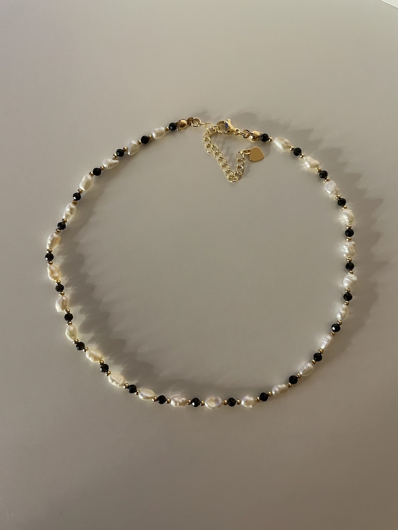 Puede incluir: Un delicado collar con perlas blancas de forma irregular intercaladas con peque&ntilde;as cuentas negras. El collar tiene un cierre y una cadena ajustable de color dorado, con un peque&ntilde;o dije en forma de coraz&oacute;n.