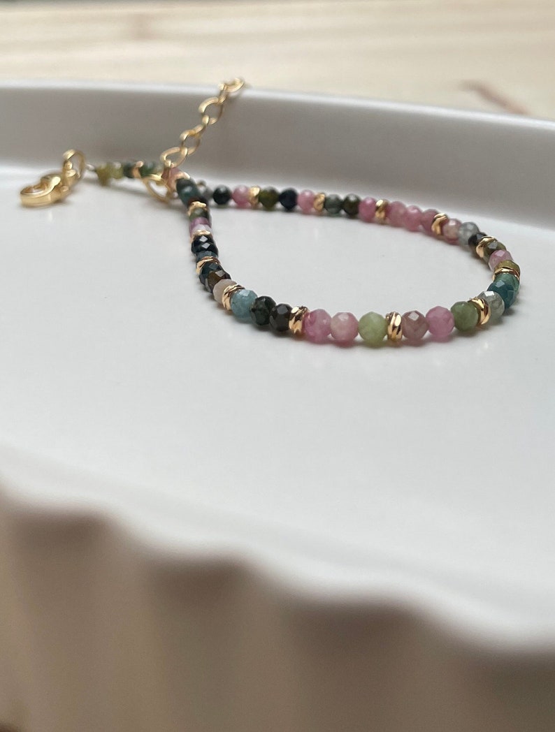 Puede incluir: Un brazalete de piedras preciosas multicolores con cuentas rosas, verdes y azules. El brazalete tiene un cierre dorado y est&aacute; enhebrado en una cadena dorada.