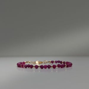 Könnte beinhalten: Ein zartes Armband mit facettierten, tiefroten Rubinperlen, die mit kleinen goldfarbenen Perlen durchsetzt sind. Das Armband wird mit einem goldfarbenen Verschluss gesichert. Der Schmuck wird auf einer hellgrauen Oberfläche präsentiert.