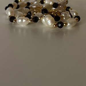 Puede incluir: Un collar con perlas blancas ovaladas, intercaladas con cuentas negras facetadas y peque&ntilde;as cuentas doradas. El collar est&aacute; dispuesto en un patr&oacute;n curvo, con las cuentas reflejando la luz.