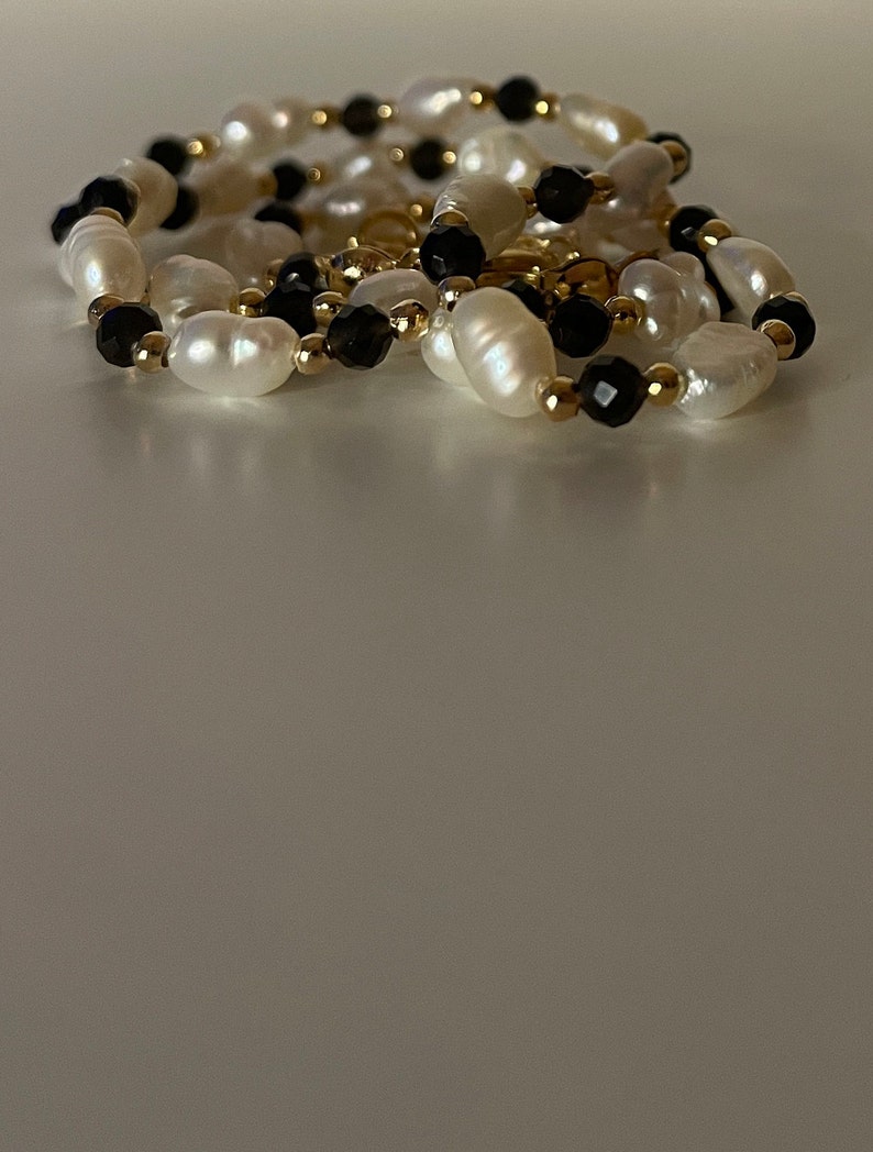 Puede incluir: Un collar con perlas blancas ovaladas, intercaladas con cuentas negras facetadas y peque&ntilde;as cuentas doradas. El collar est&aacute; dispuesto en un patr&oacute;n curvo, con las cuentas reflejando la luz.