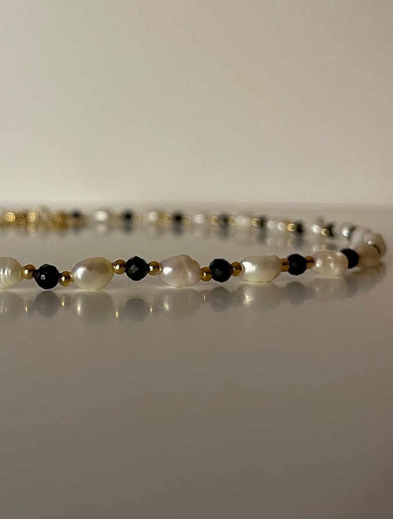 Puede incluir: Un delicado collar con perlas blancas de forma irregular, peque&ntilde;as cuentas negras y detalles dorados. El collar se muestra sobre un fondo neutro, resaltando su elegante dise&ntilde;o. La longitud del collar es de aproximadamente 40 cm.