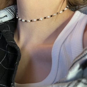 Puede incluir: Un delicado collar gargantilla con una hebra de perlas blancas, intercaladas con peque&ntilde;as cuentas negras y detalles dorados. El collar se lleva alrededor del cuello, creando un accesorio sutil y elegante. La joya est&aacute; sobre un fondo neutro.