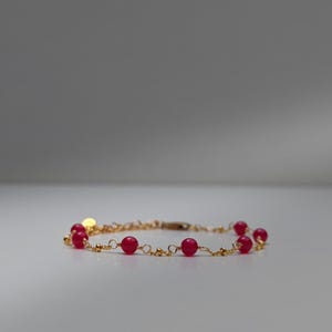 Op de afbeelding: Een delicate goudkleurige armband met kleine, ronde, rode kralen. De kralen zijn gelijkmatig verdeeld over de gouden ketting. Een kleine, ronde gouden bedel is aan de armband bevestigd.