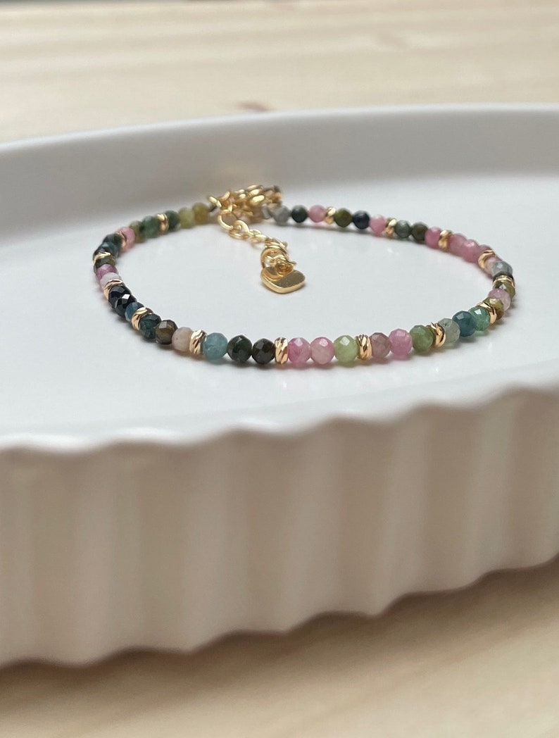 Puede incluir: Un delicado brazalete hecho con peque&ntilde;as cuentas de piedras preciosas multicolores. Las cuentas son una mezcla de rosa, verde y azul, y est&aacute;n intercaladas con detalles dorados. El brazalete tiene un dije en forma de coraz&oacute;n.