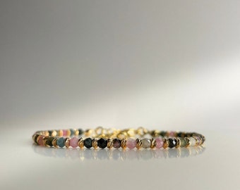 May include: A gold bracelet featuring a fine chain and small, multicoloured gemstones. The gemstones are pink, green, and black.