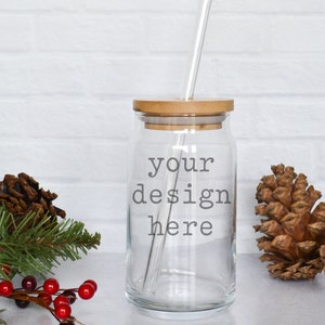 May include: A clear glass jar with a bamboo lid and a straw. The jar has the text "your design here" printed on it. The jar is sitting on a white surface with pine cones and a sprig of evergreen.
