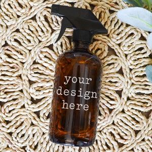May include: A brown glass spray bottle with a black trigger sprayer. The bottle has a white label with the text "your design here". The bottle is sitting on a woven straw mat.