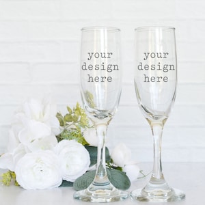 May include: Two clear champagne flutes with the text "your design here" are displayed. A bouquet of white flowers and greenery is arranged to the left of the flutes. The background is a white brick wall.