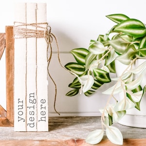 May include: A stack of three white books with the text "your design here" printed on the side. The books are tied together with twine and are sitting on a wooden shelf next to a potted plant with green and white leaves.