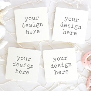 May include: Four white square coasters with the text "your design here" printed on each. The coasters are arranged in a square shape on a white surface with a white fishing net and seashells.