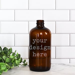 May include: A brown glass bottle with a white label that says "your design here" on a white tiled background with green foliage.