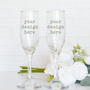 May include: Two clear glass champagne flutes with the text "your design here" on each. The flutes are sitting on a white surface with a bouquet of white flowers and green leaves.