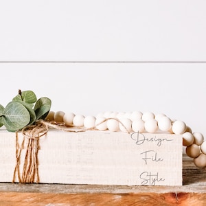 May include: A white wooden block with the words "Design File Style" written on it. The block is decorated with a sprig of greenery and twine. A strand of white wooden beads is draped over the block.