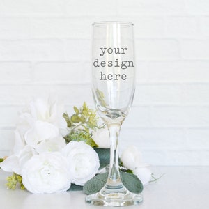 May include: A clear glass champagne flute with the text "your design here" on the side. The flute is sitting on a white surface next to a bouquet of white flowers.