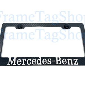 May include: Black license plate frame with a carbon fiber pattern and the words "Mercedes-Benz" in white. The frame has two black screw caps at the top. A stylish car accessory.
