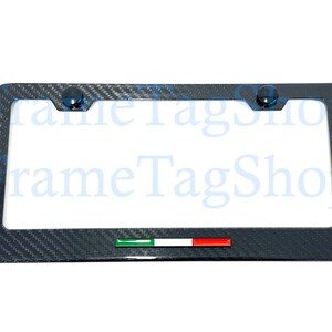 May include: A carbon fiber license plate frame with a rectangular shape. The frame has a dark gray woven pattern and features two dark blue screw caps. An Italian flag emblem is at the bottom.