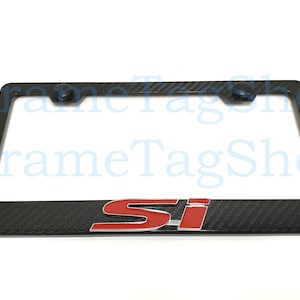 May include: Black license plate frame with a carbon fiber pattern and the red letters "Si". The frame has two black mounting caps at the top. The product is for automotive use.