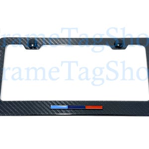 May include: A black license plate frame with a carbon fiber weave pattern. The frame has a rectangular shape with rounded corners and includes a blue, dark blue, and red emblem. Two black fasteners are visible at the top.
