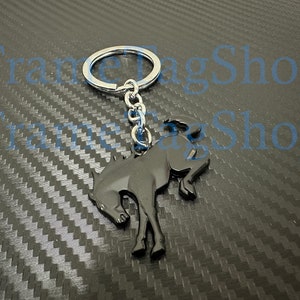 May include: Black metal keychain featuring a stylized horse silhouette. The keychain has a silver ring and chain. The horse design is a dark, glossy black. The background is a textured, dark gray surface.