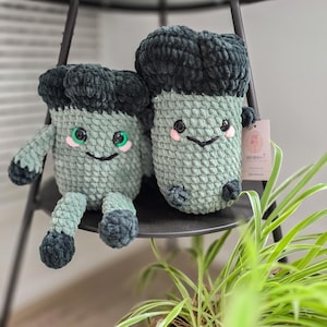 May include: Two crocheted plush toys shaped like green monsters with black hair. The monsters have smiling faces with green eyes and pink cheeks. The toys are sitting on a black shelf.
