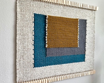 May include: A woven wall hanging with a geometric design featuring squares and rectangles in shades of teal, gray, and gold. The hanging is framed with a wooden dowel.