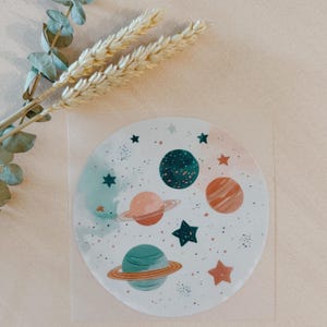 Könnte beinhalten: Ein runder, weißer Untersetzer mit einer farbenfrohen Illustration von Planeten und Sternen. Die Planeten sind in Orange, Türkis und Grün gehalten, mit Sternakzenten. Getrocknete Weizen- und Eukalyptuszweige sind zu sehen.