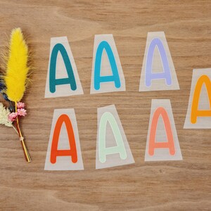 Könnte beinhalten: Eine Sammlung farbenfroher "A"-Buchstabenaufkleber in verschiedenen Farbtönen, darunter Türkis, Blau, Lila, Orange und Grün. Ein kleiner Strauß Trockenblumen mit gelben, rosa und blauen Akzenten ist ebenfalls vorhanden.