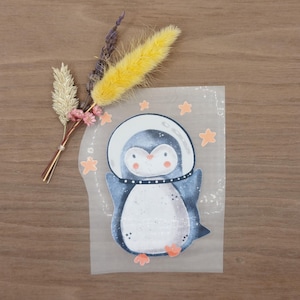 Könnte beinhalten: Eine skurrile Illustration eines Pinguins mit Weltraumhelm, blau-weißem Körper, orangefarbenen Füßen und einem gepunkteten Kragen. Der Pinguin ist von orangefarbenen Sternen auf transparentem Hintergrund umgeben. Getrocknete Blumen in Gelb, Beige und Lila.